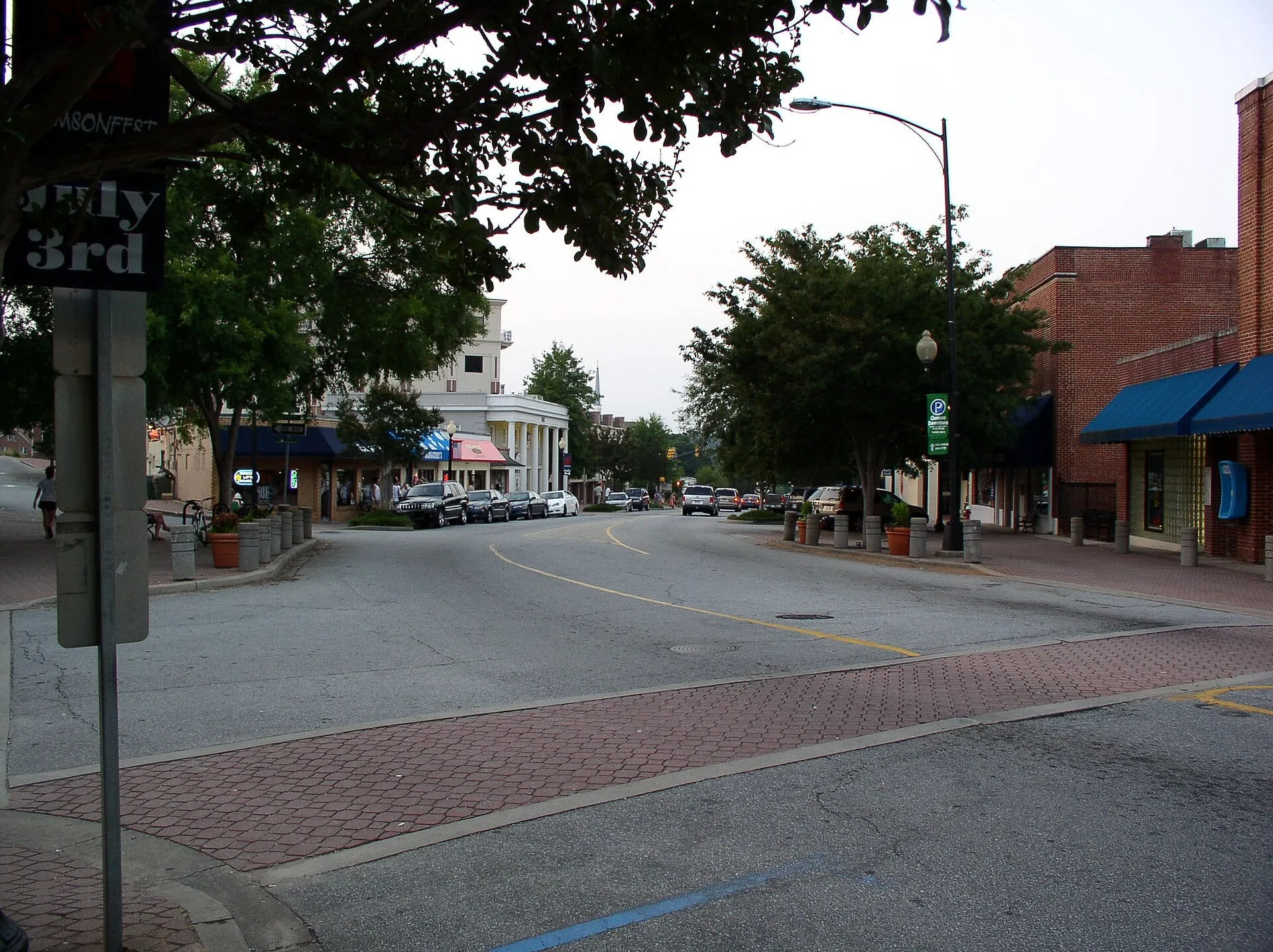 College Avenue in downtown Clemson, with brick storefronts and blue awnings on the right and cars parked under shade trees